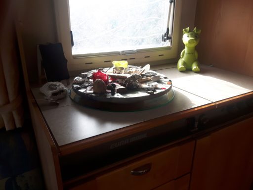 Runder Tisch mit Spielzeug, Sammelobjekten und grüner Figur, vor Fenster.